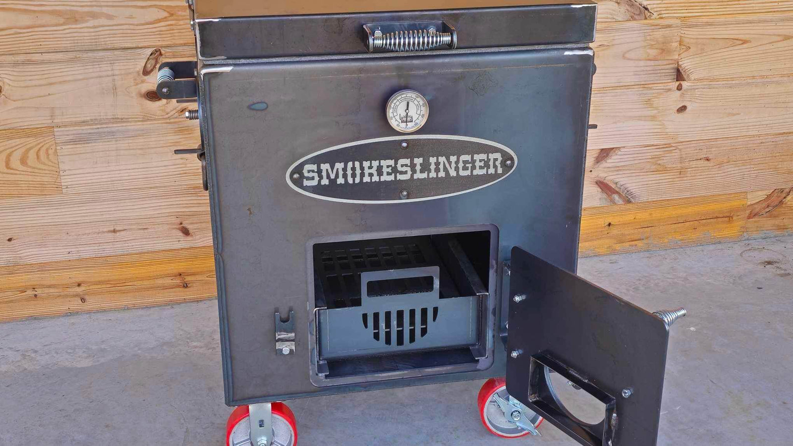 image of the top view of a smokeslinger smoker with the fire box door open sitting on conrete with a natural stained wood wall in the bacground