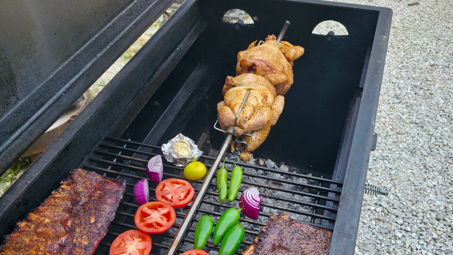 Two chickens turning on a rotisserie spit above the grate of a Full Size Smokeslinger, with ribs and vegetables cooking below