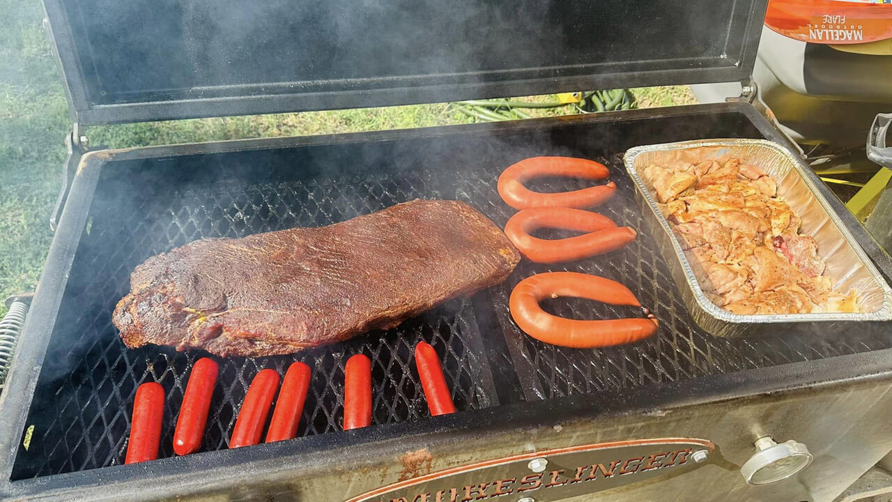 image of the top view of a smokeslinger smoker sitting outside with the top open and a slab of ribs, chicken, and sausage cooking
